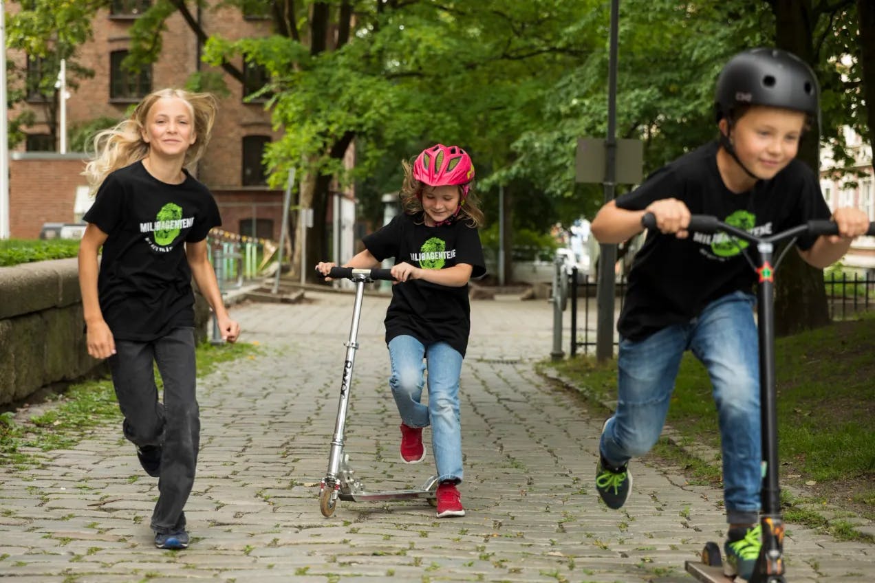 Tre miljøagenter i en skolegård. Jenta til venstre i bildet løper, og de to andre sparker på sparkesykkel. Fotografi.