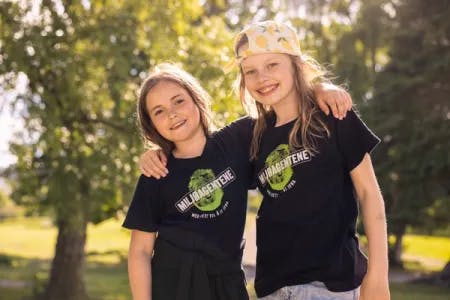 To jenter står ved siden av hverandre og holder hverandre om skuldrene mens de smiler. De har på seg svarte t-skjorter med Miljøagentenes logo. Jenta til høyre har på en caps bak frem. De står i en park og det er sommer.