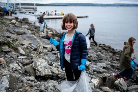 En jente står i fjæra, hun har på seg blå hansker og holder en søppelpose. I bakgrunnen er det flere miljøagenter som også rydder søppel.Fotografi.