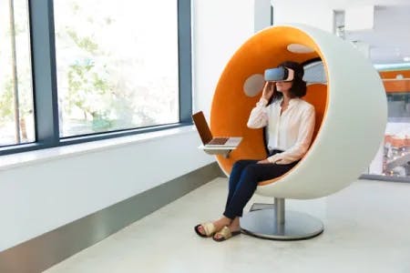 En dame sitter i en hvit rund stol, med orange for. Hun har på seg VR briller. Fotografi.