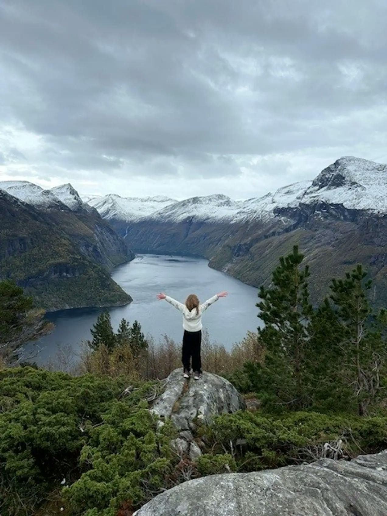 Jente på en stein med armene over hodet. Ser på utsikten, på vann og fjell. Fotografi.