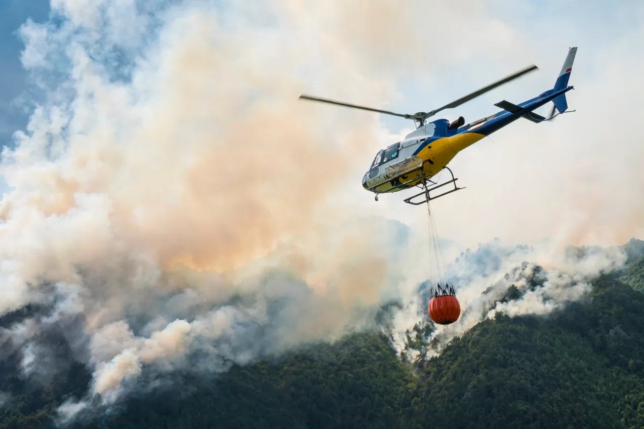 Et helikopter med vannbøtte forsøker å slukke skogbrann. Fotografi.