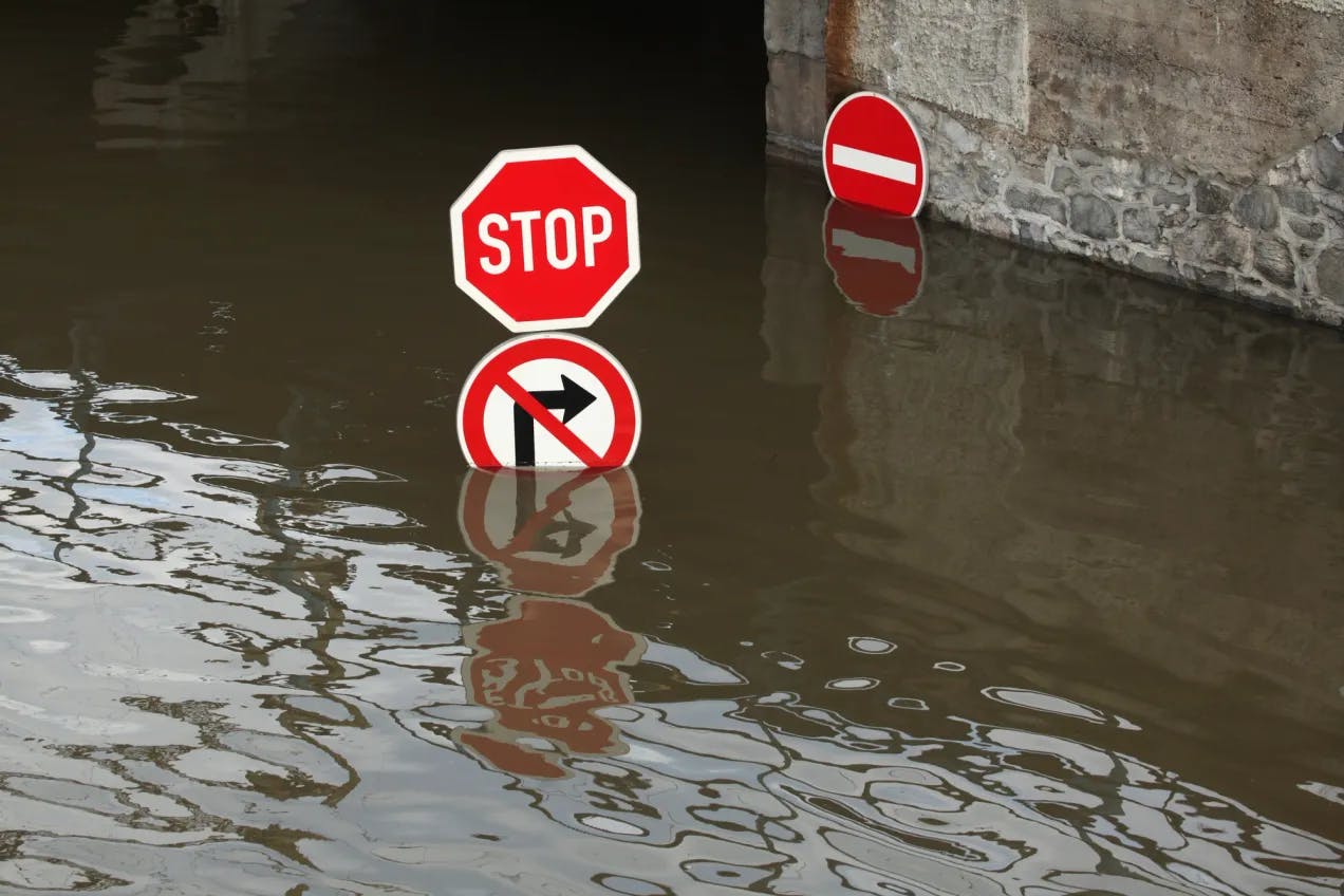 En oversvømt vei, hvor vannet når helt opp til veiskiltene. Det er tre skilt i bildet: stopp-skilt, forbudt å kjøre til høyre-skilt og innkjøring forbudt-skilt. Fotografi.