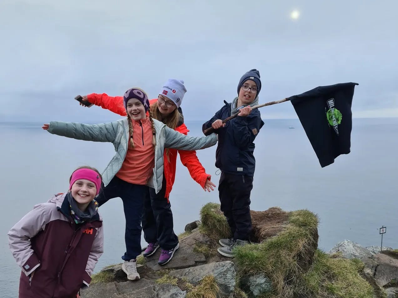 Flere miljøagenter som står på et fjell. De holder et miljøagentflagg. Vi ser havet i bakgrunnen.