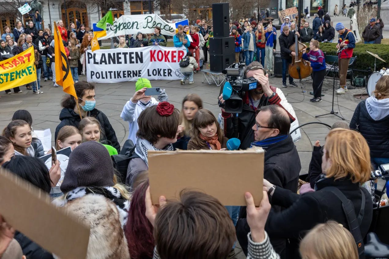 Flere titalls personer samlet på Eidsvolls plass foran stortinget. Espen Barth Eide blir intervjuet av NRK, mange har plakater og flagg. Et band synger og spiller, en har mikrofon og en har kontrabass. På det største banneret står det "Planeten koker. Politikerne loker"