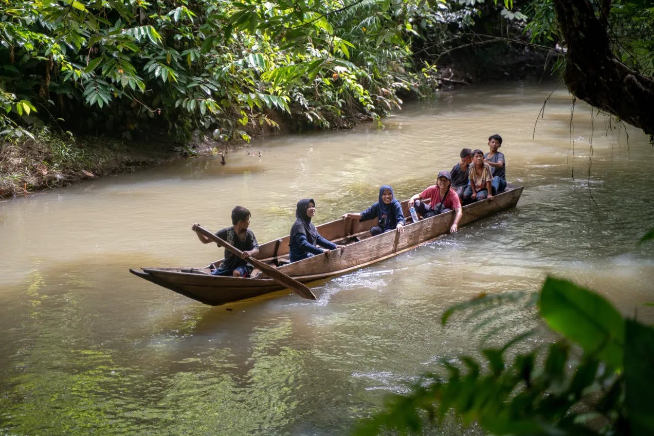 En brun elv med tett, grønn vegetasjon lands elvebreddene. Midt på elva er en tynn, brun båt av tre med 7 barn. Gutten foran holder en åre av tre som han ror med.