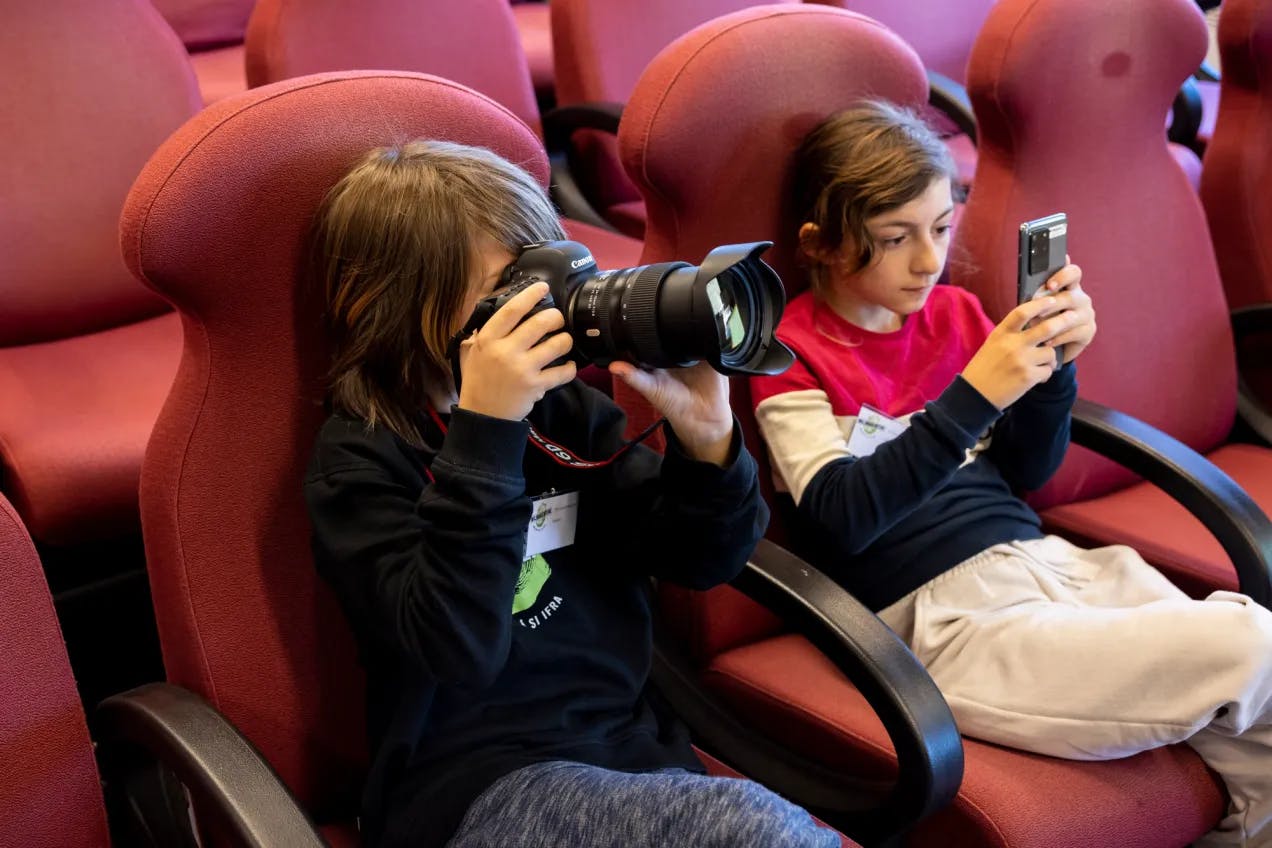 To miljøagenter sitter i lyserøde kinoseter. En holder et speilreflekskamera, den andre miljøagenten holder en mobiltelefon. Begge tar bilder av noe utenfor bildet.