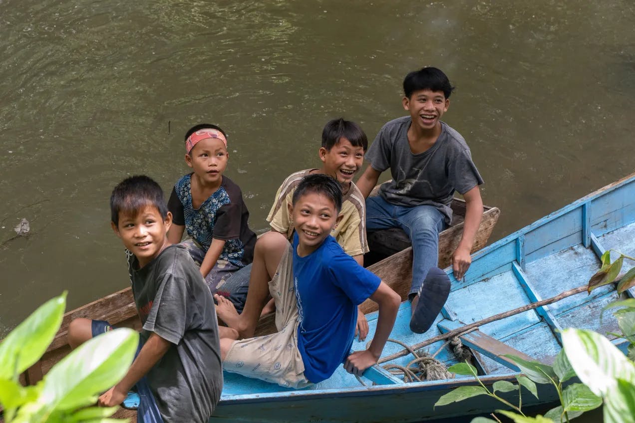 Fem smilende barn sitter i en liten trebåt i en brun elv. Bilde tatt ovenfra og de ser oppover. I forgrunnen ser vi grønne blader.