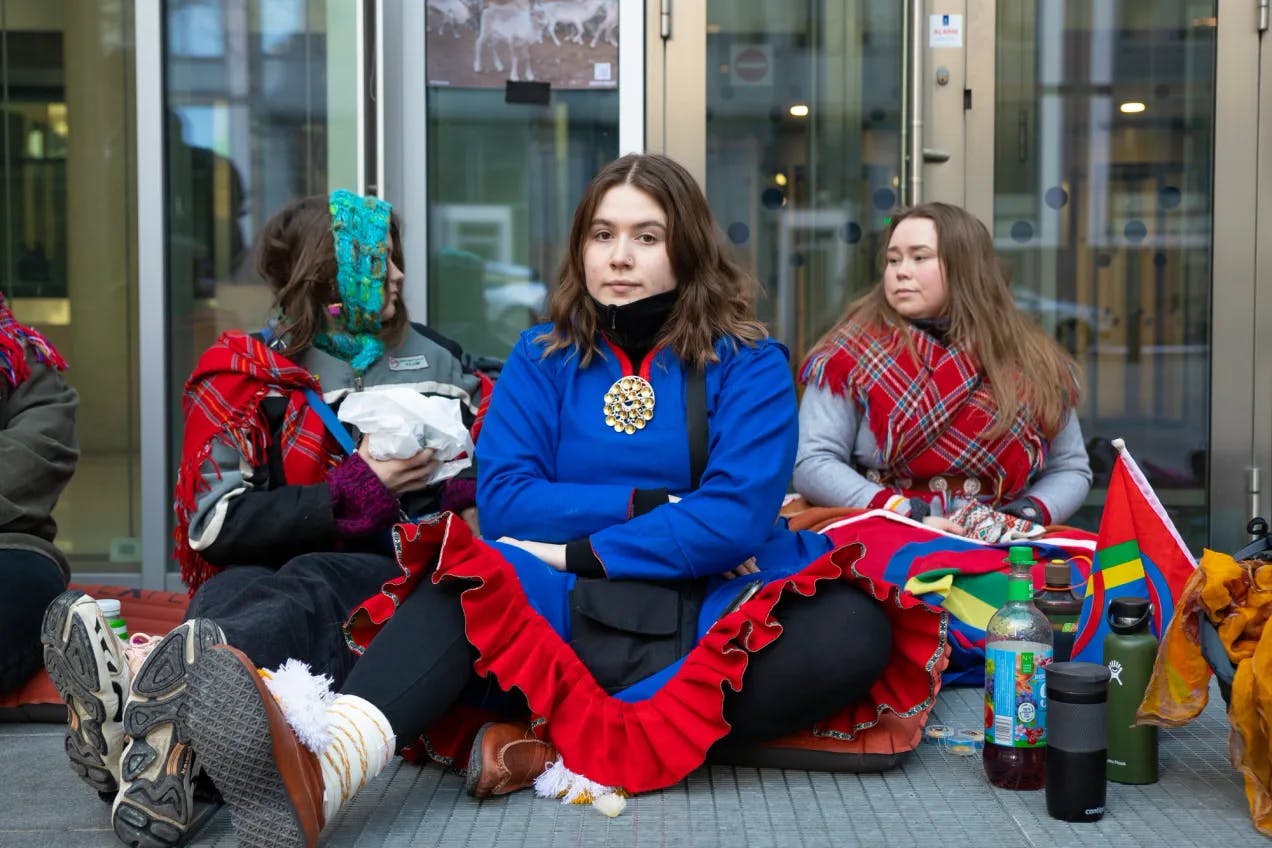 Tre jenter sitter på bakken foran en dør. Jenta i midten har på seg samisk kofte og sølje. Jentene ved siden av har på seg samiske sjal. Foran står en saftflaske og to termoser, og det samiske flagget.