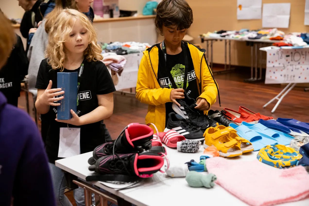 En gutt og en jente står og ser på et bord med sko og klær. De er på byttemarked og lurer på hva de skal velge. Begge har sorte miljøagent t-skjorter og gutten har også en gul genser.