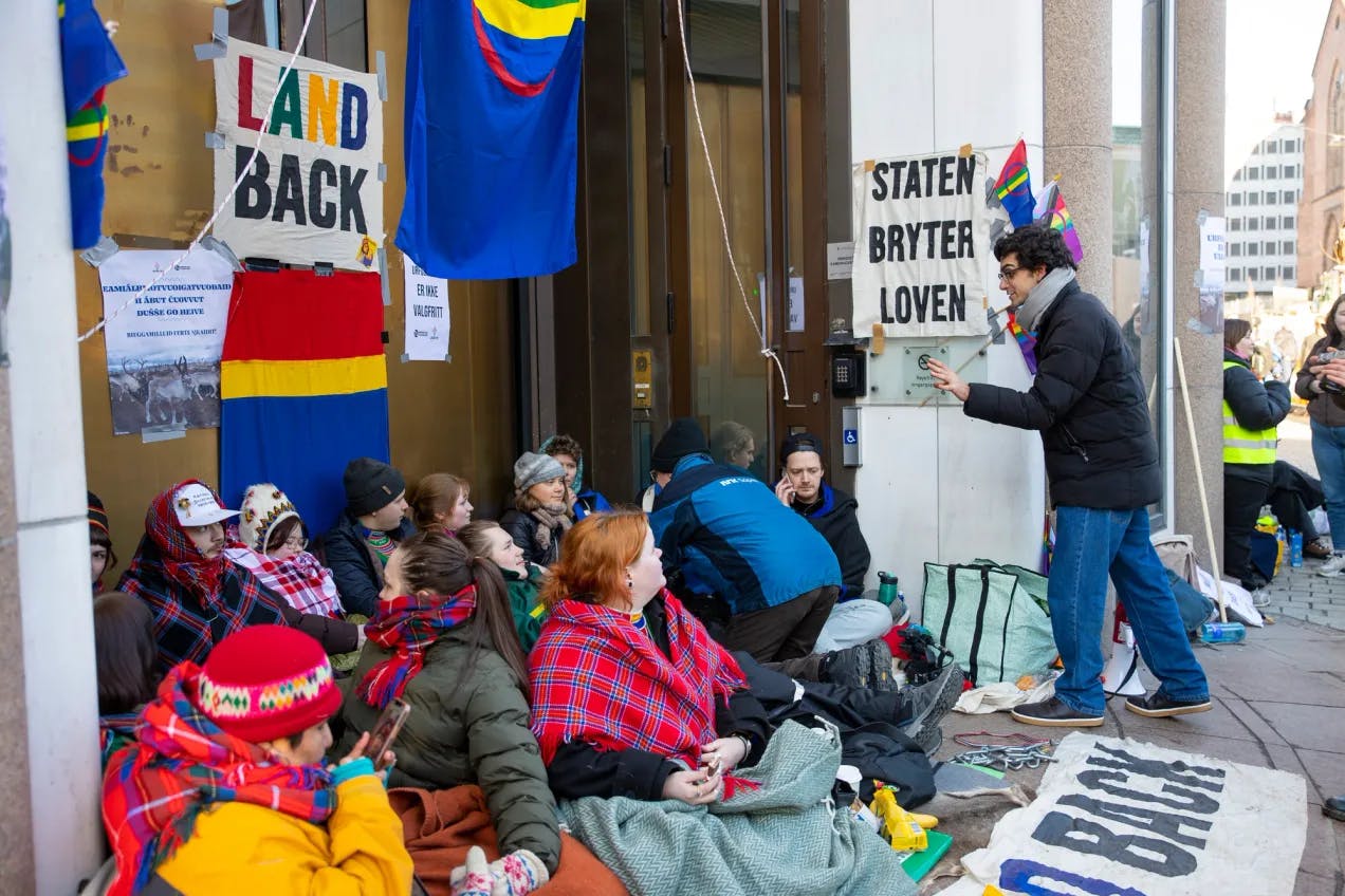 Cirka 12 ungdommer sitter foran dørene til olje- og energidepartementet. De har på seg varme jakker, luer og noen har på seg samiske sjal. Bak henger samiske flagg, og bannere hvor det står "Land back" og "Staten bryter loven"