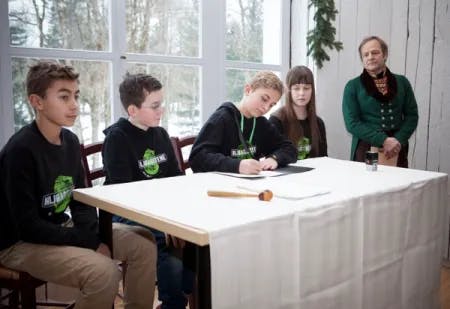 Fire barn med Miljøagentene-klær signerer et dokument i Rikssalen på Eidsvoll i forbindelse med grunnleggingen av Barnas klimapanel i 2017. En voksen med klær inspirert av klærne til Eidsvoldmennene følger med ved siden av dem