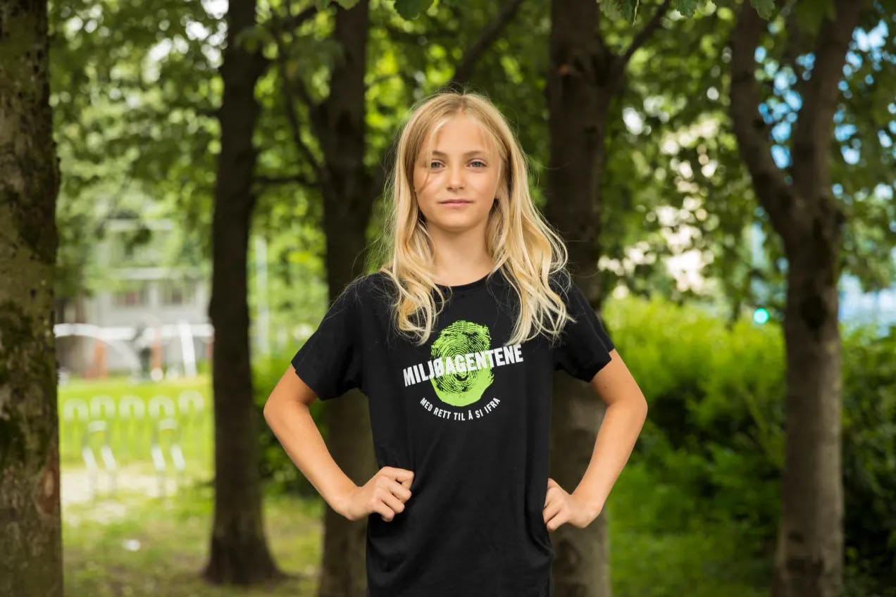 En jente med blondt hår står med armene i siden. Hun har på seg en svart Tskjorte med Miljøagentene-logo på. I bakgrunnen ser man løvtrær og skimter et hus i bakgrunnen.
